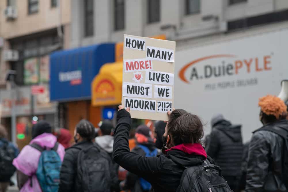 Protestor holding a sign that says "How Many More Lives Must We Mourn?"